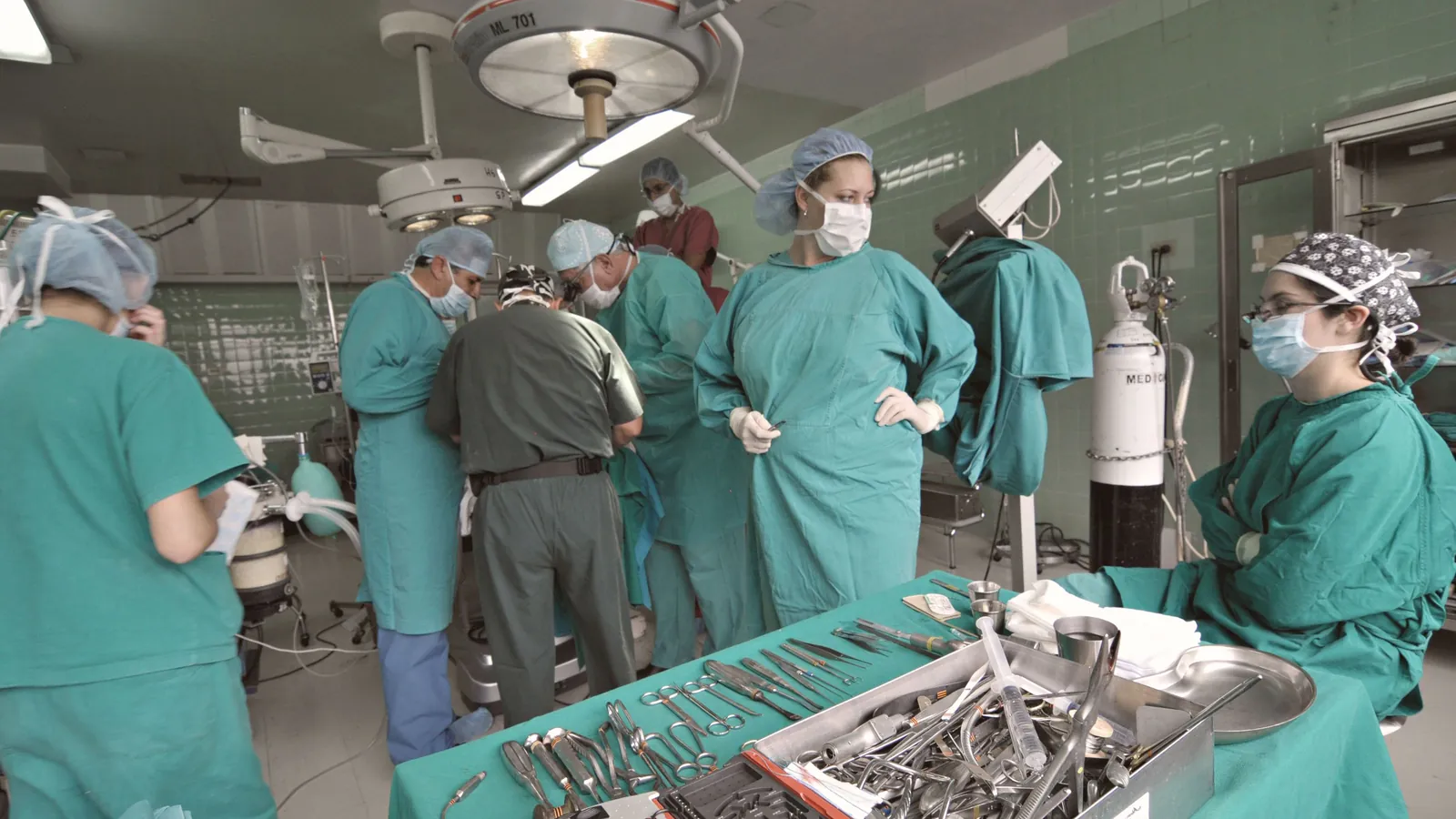 A surgical team in green scrubs at work in an operating room, each person at a different station