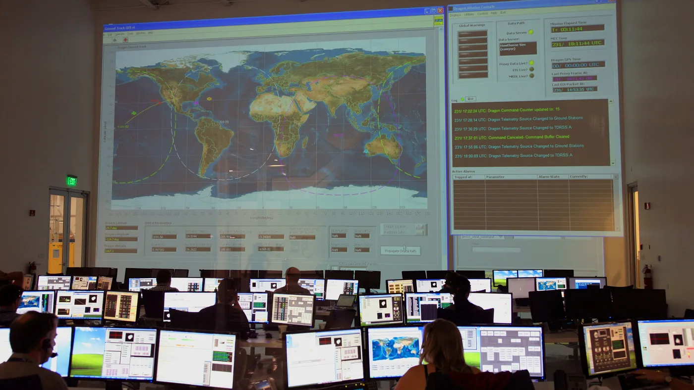 Rows of operators at computer screens in a control room facing a large wall display with a world map