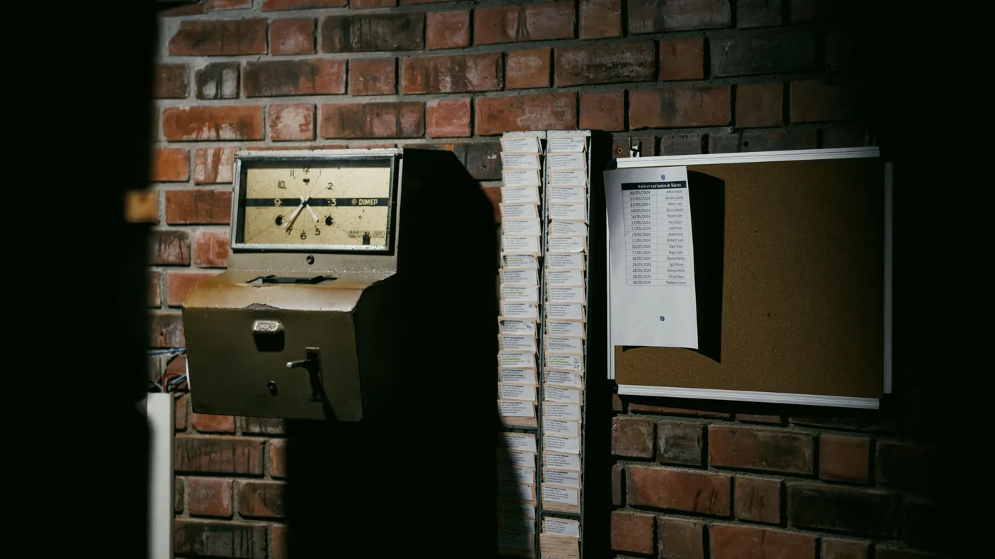 A vintage factory time clock mounted beside a rack of punch cards on a brick wall