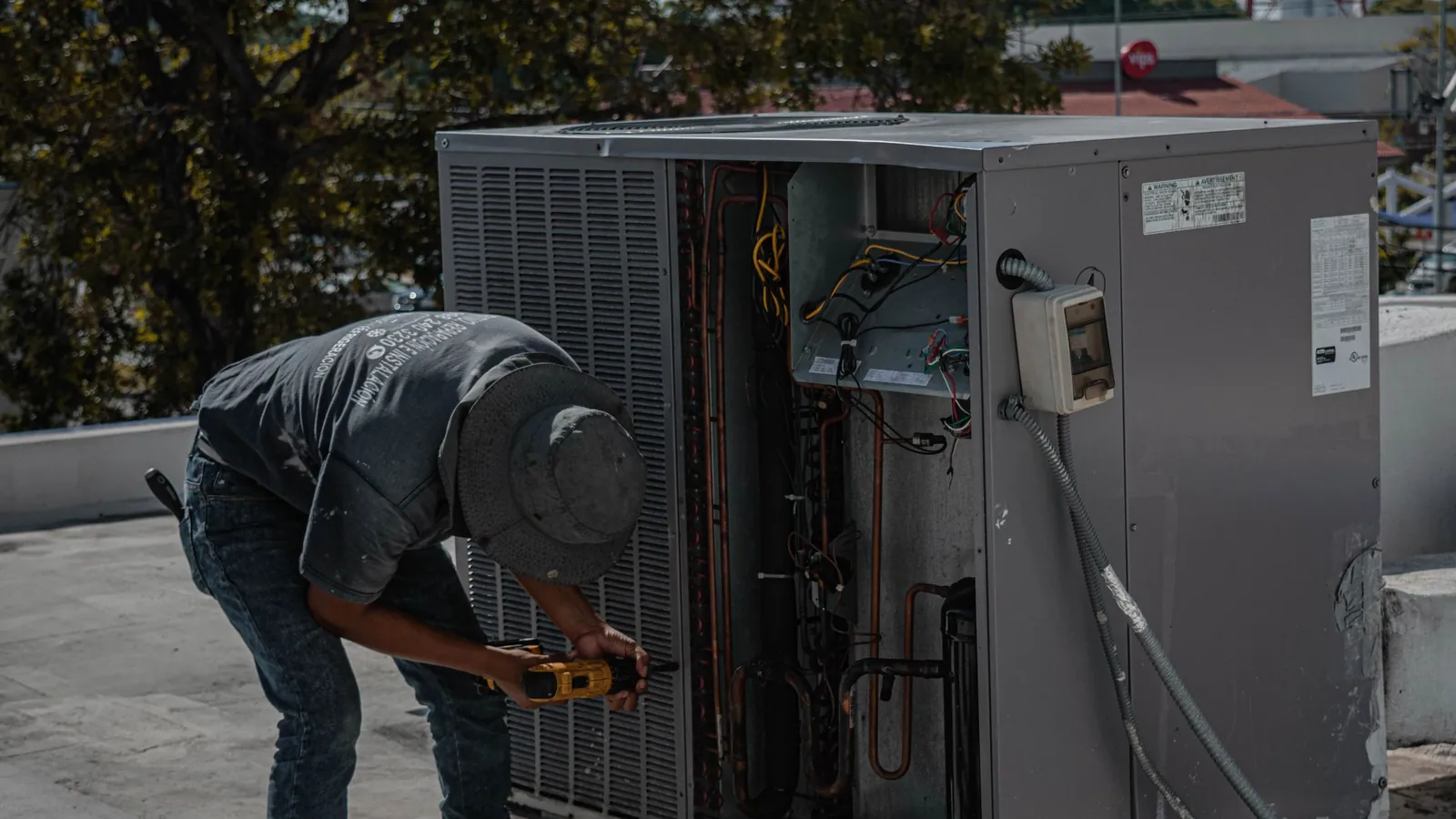 A technician in a bucket hat working inside an open rooftop air conditioning unit with a power drill