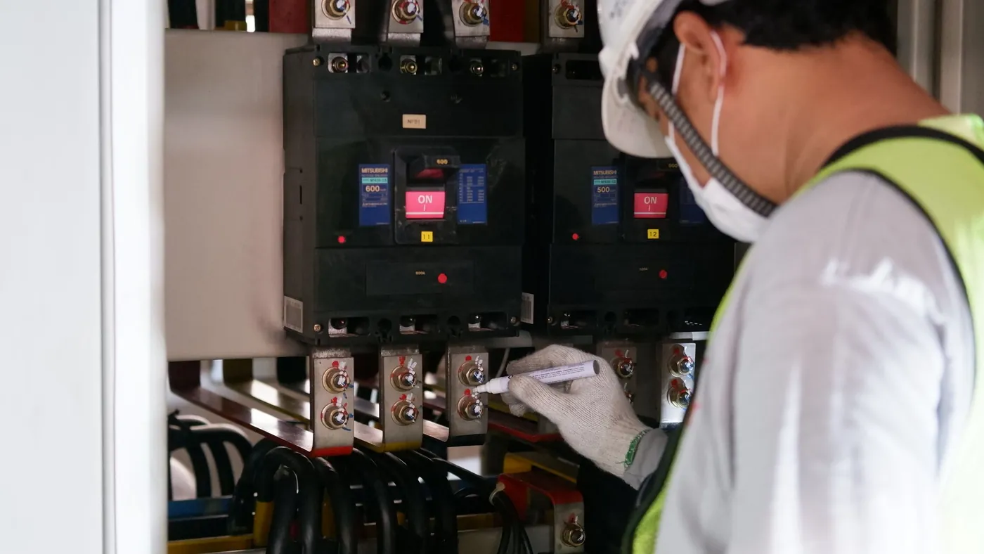 A technician in a hard hat, mask and high-visibility vest marking a bolt on an industrial electrical panel with a paint pen