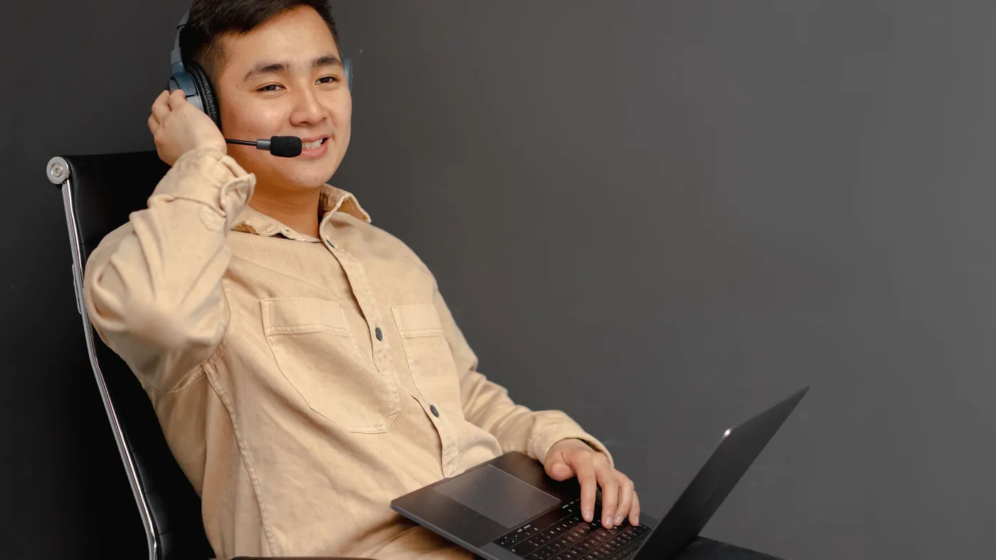 A smiling customer service agent in a headset sitting back in an office chair with a laptop on his lap