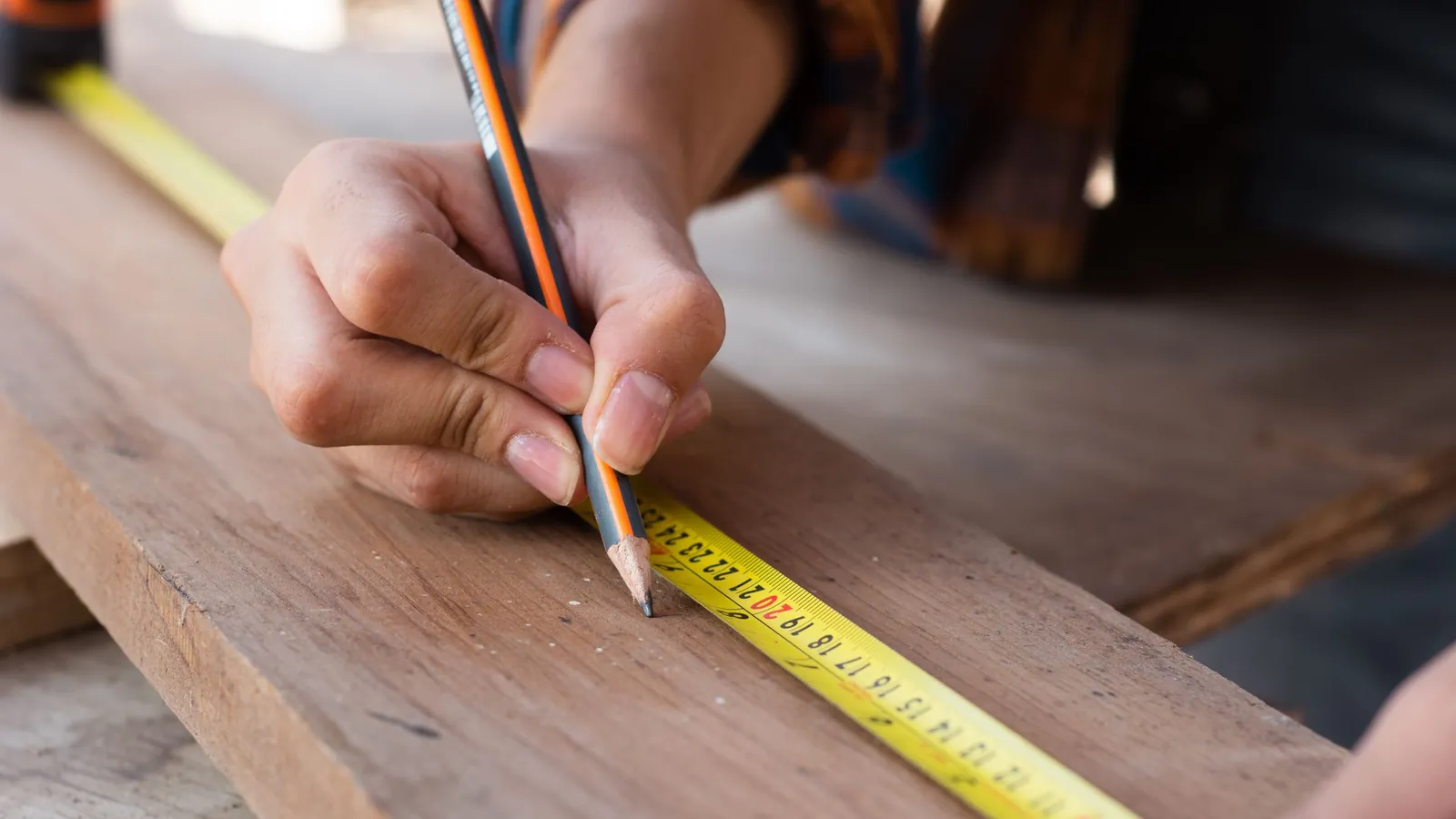 A hand marking a wooden plank with a pencil against a yellow tape measure