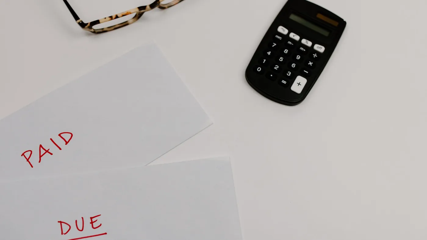 Two sheets of paper marked paid and due in red ink beside a calculator and a pair of glasses on a white desk