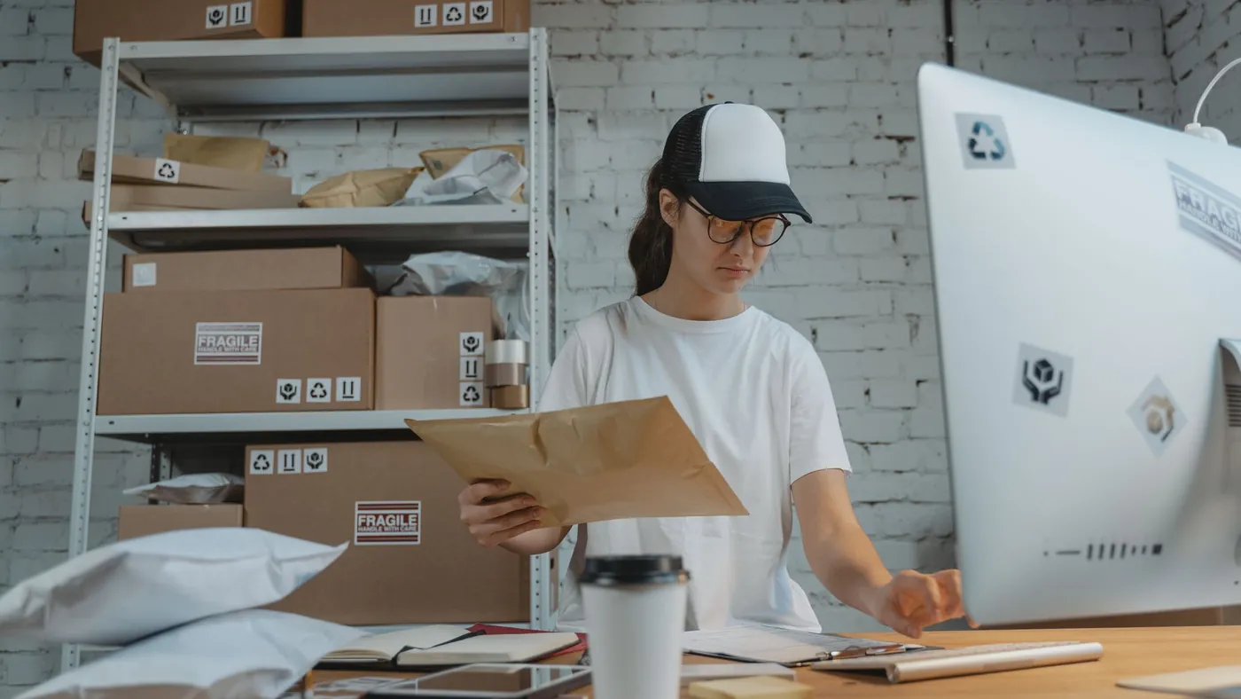 A woman in a cap and glasses reading a padded envelope at a desk in a stockroom lined with boxes