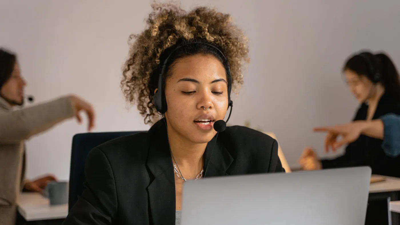 A customer support agent wearing a headset speaks while working on a laptop in an office