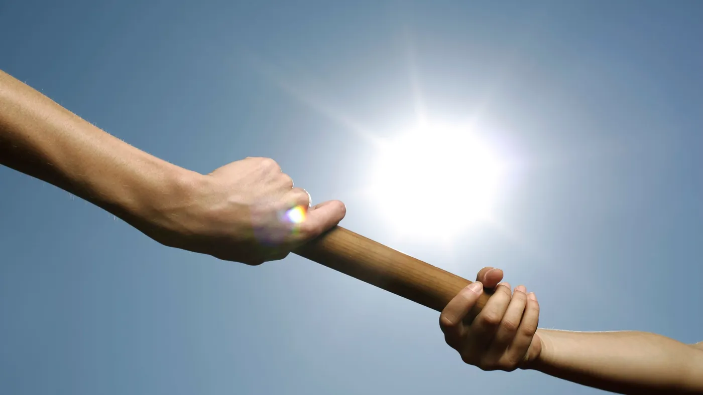 Two hands passing a wooden relay baton against a bright blue sky