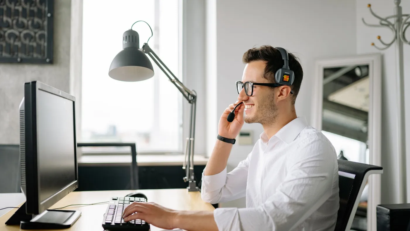 A man in a headset smiling at a customer while typing at a desktop computer in a bright office
