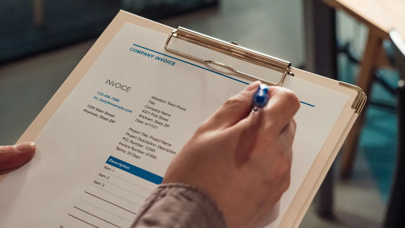 A hand holding a pen over a printed company invoice clipped to a clipboard