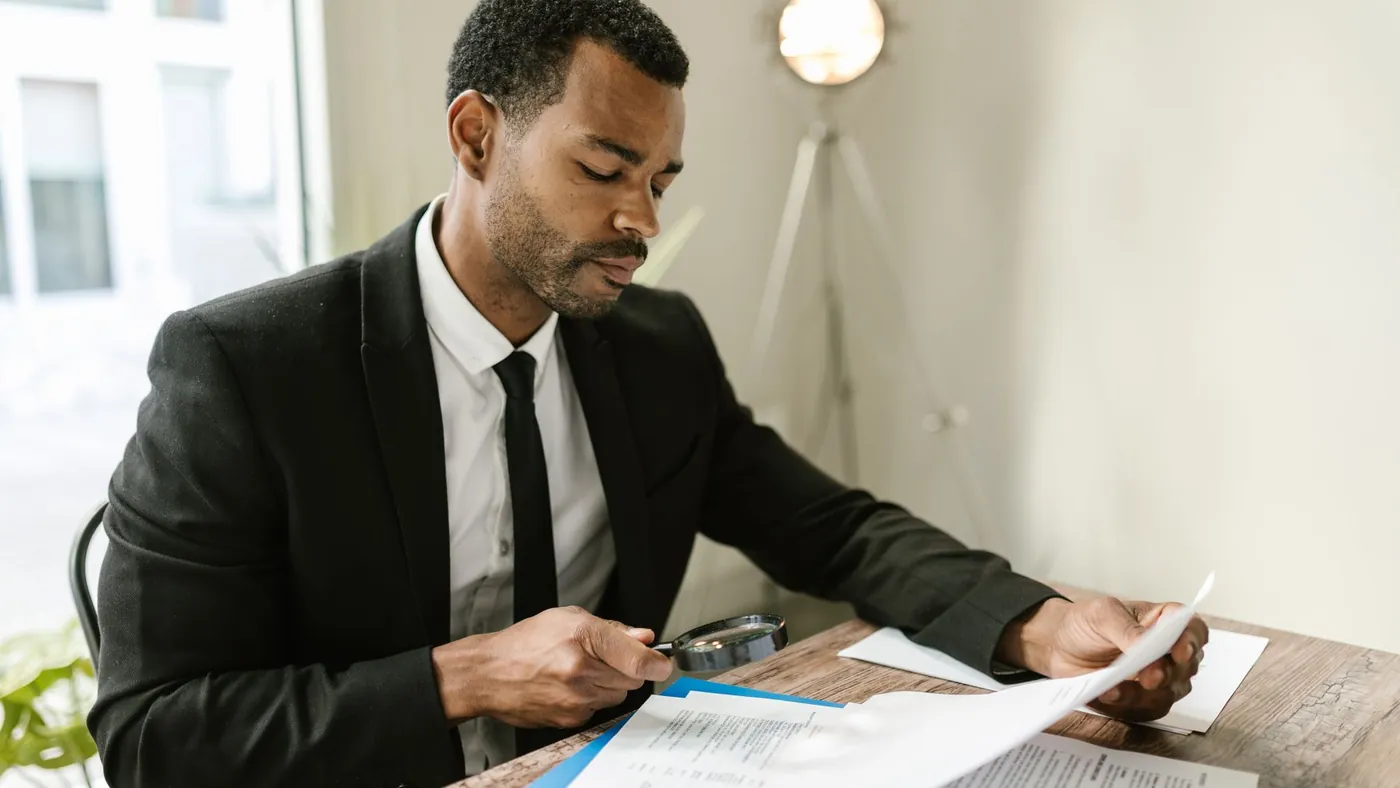 A man in a suit examining a printed document with a magnifying glass at his desk