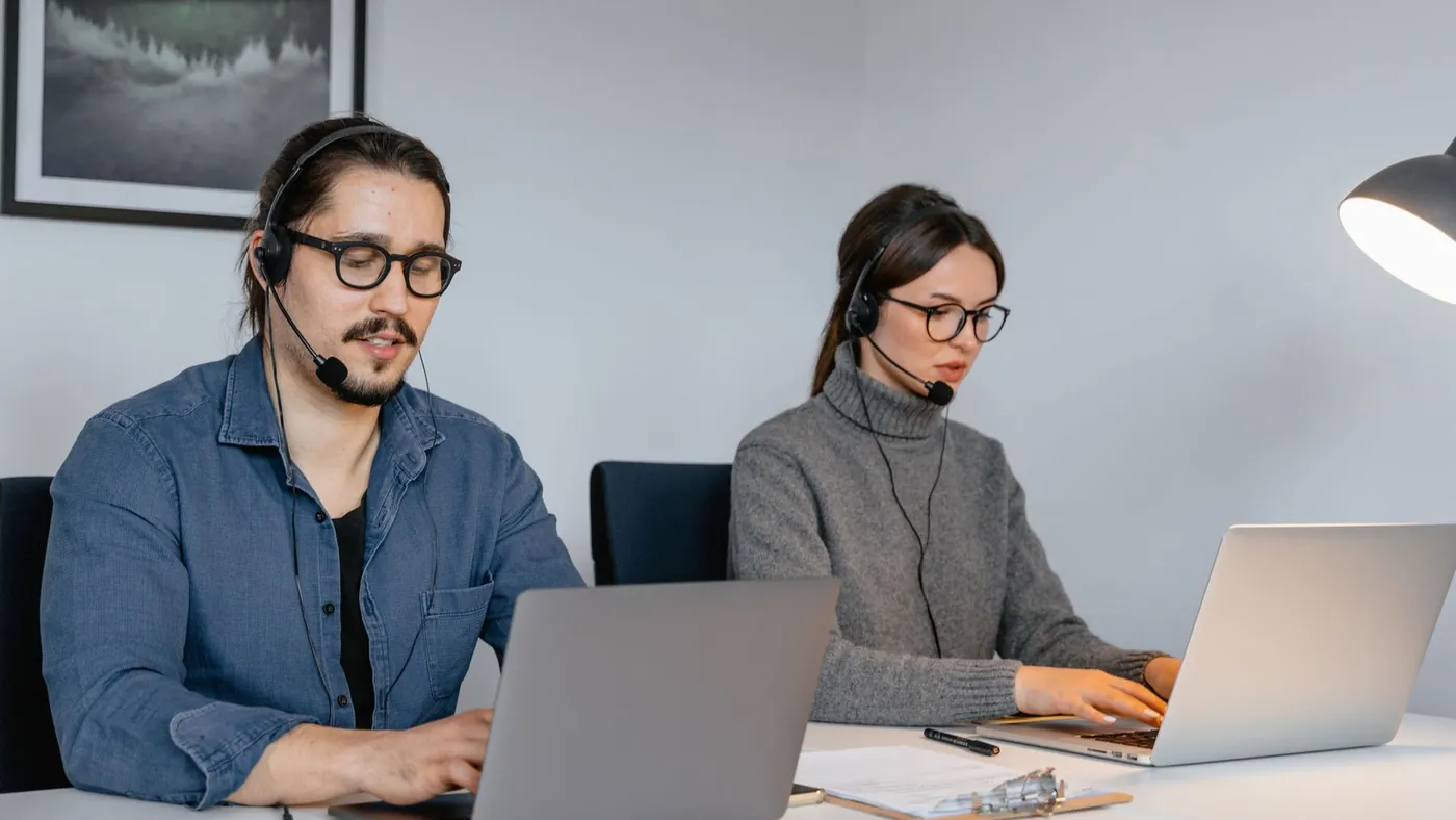 Two customer support agents wearing headsets working on laptops at a shared desk