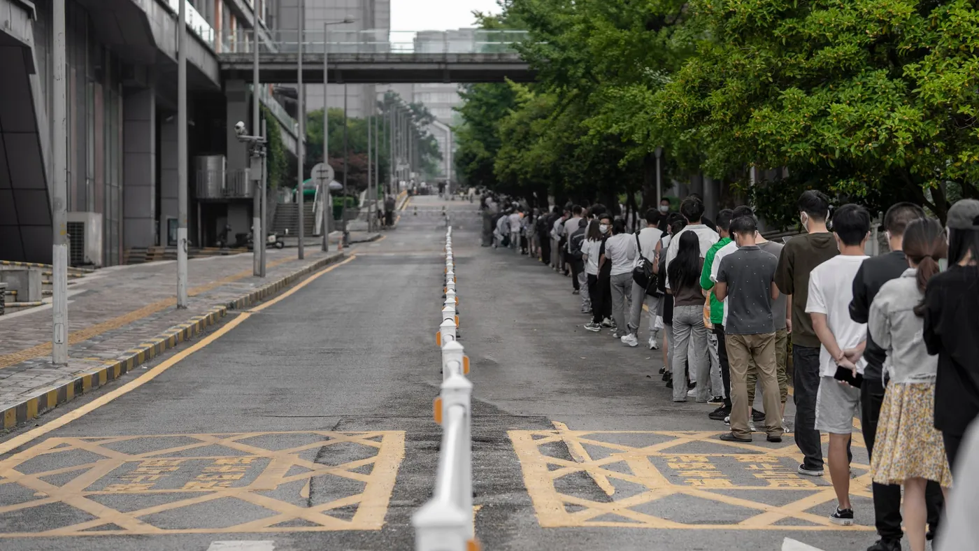 A long line of people waiting along the edge of a tree-lined city street