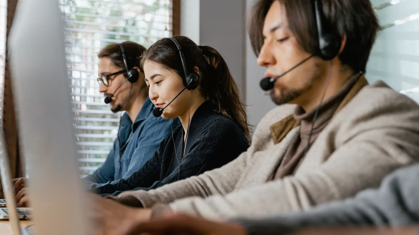 Three support agents wearing headsets work at computers in a row in a bright office