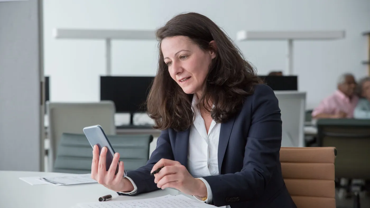 A woman in a blazer at an office desk reading her phone with papers and a pen in front of her