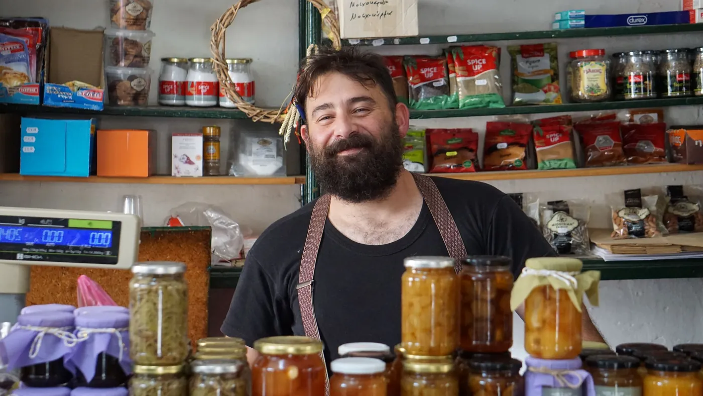 A smiling shopkeeper standing behind wooden shelves lined with jars of homemade preserves