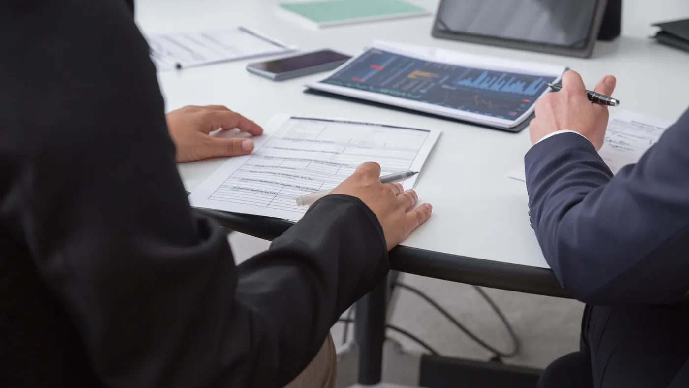 Two people at a meeting table reviewing a printed form and charts with pens in hand