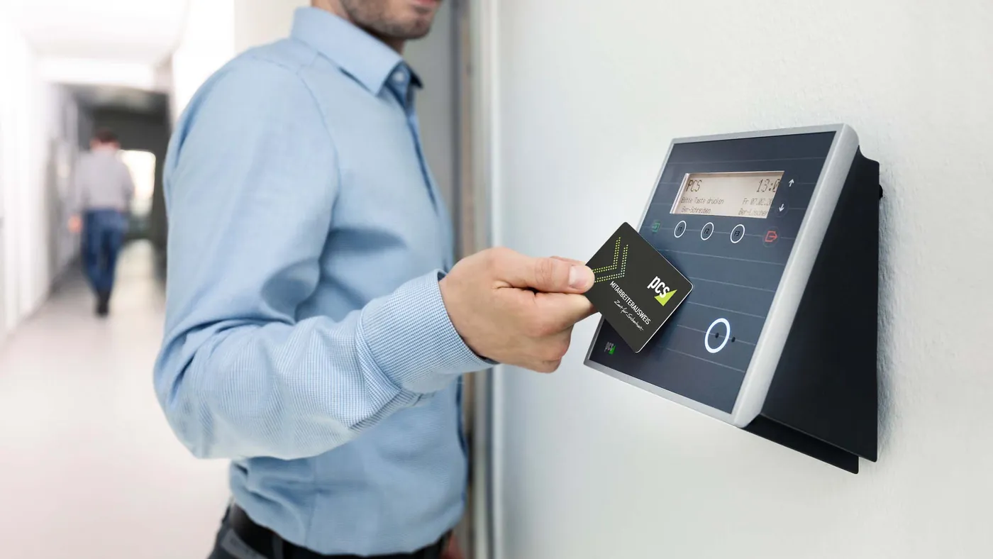 A person in a blue shirt holding an employee badge against a wall-mounted card reader