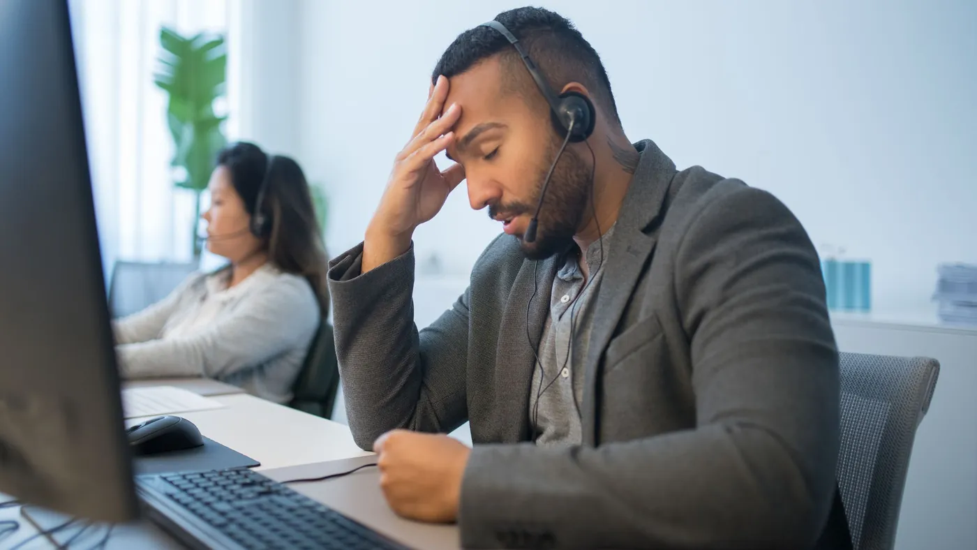 A tired call centre worker with a headset resting his head in his hand at his desk