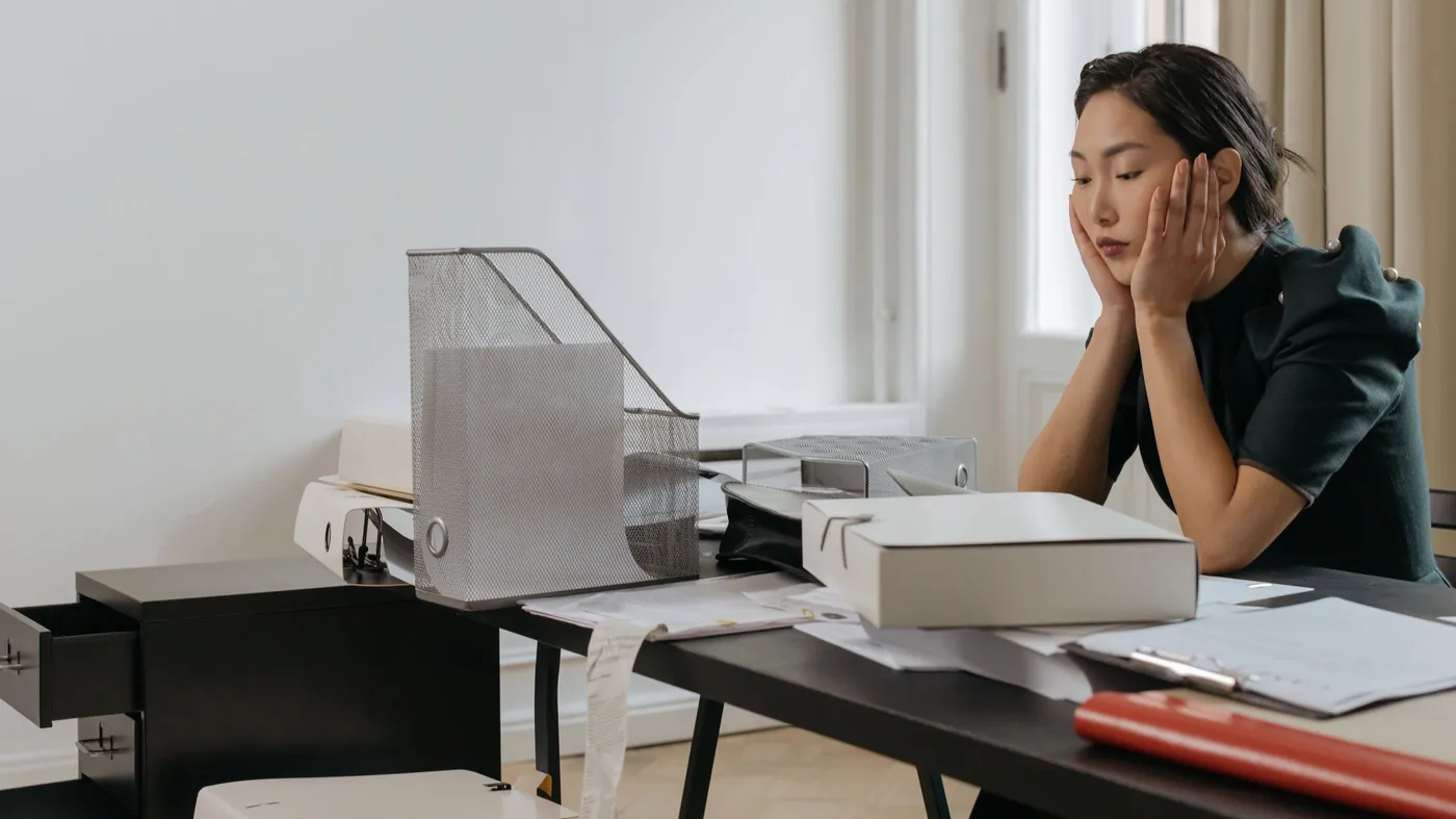 A woman at an office desk with her hands on her cheeks, surrounded by paperwork