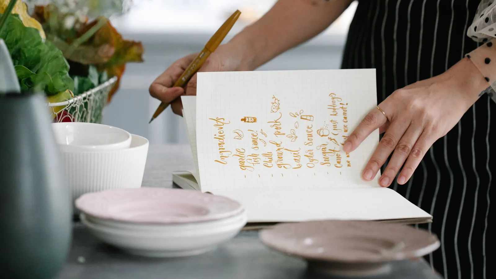 A person in an apron writing a handwritten recipe into an open notebook on a kitchen counter