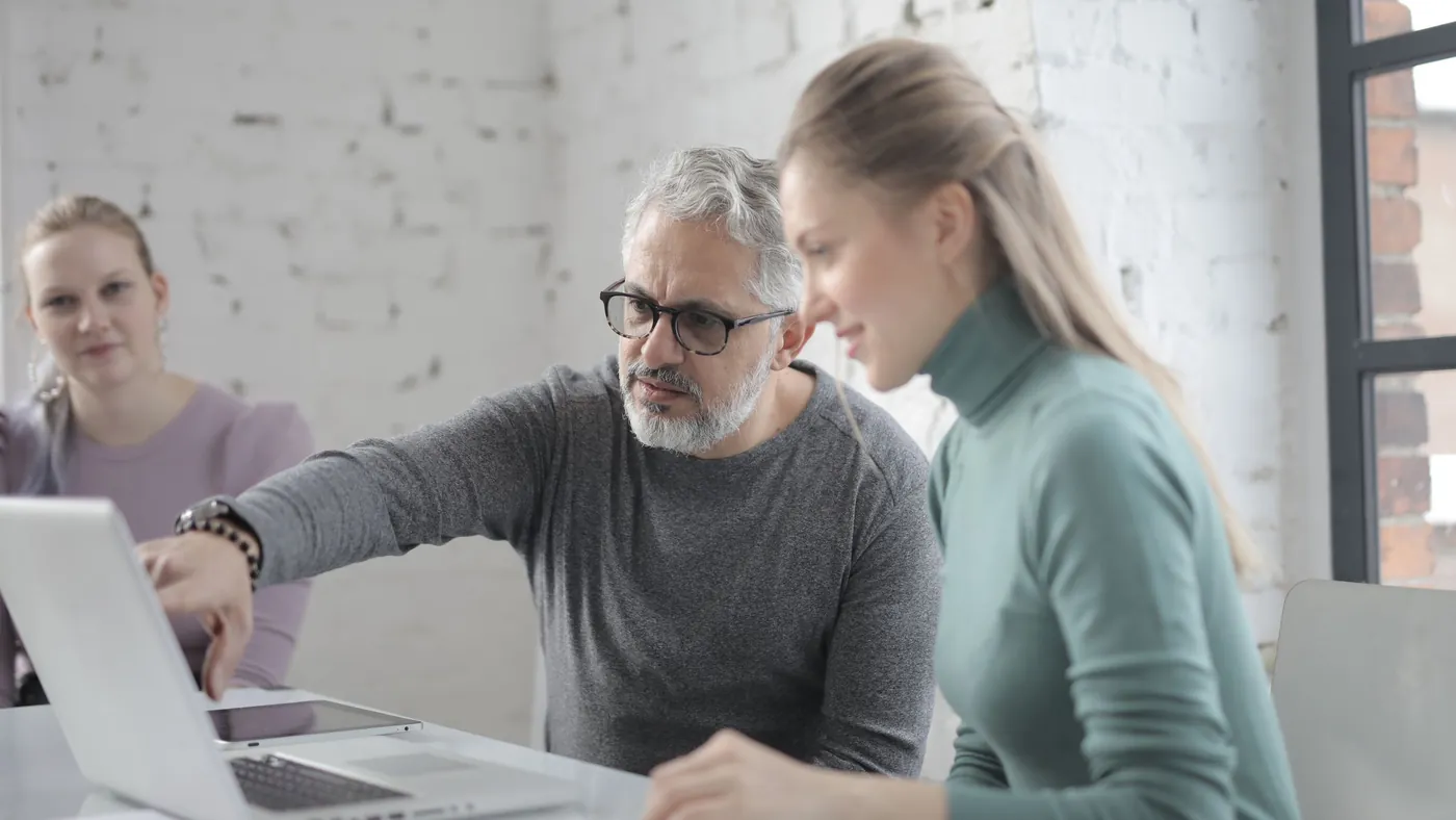 An older colleague pointing at a laptop screen while explaining something to a younger coworker at a shared desk