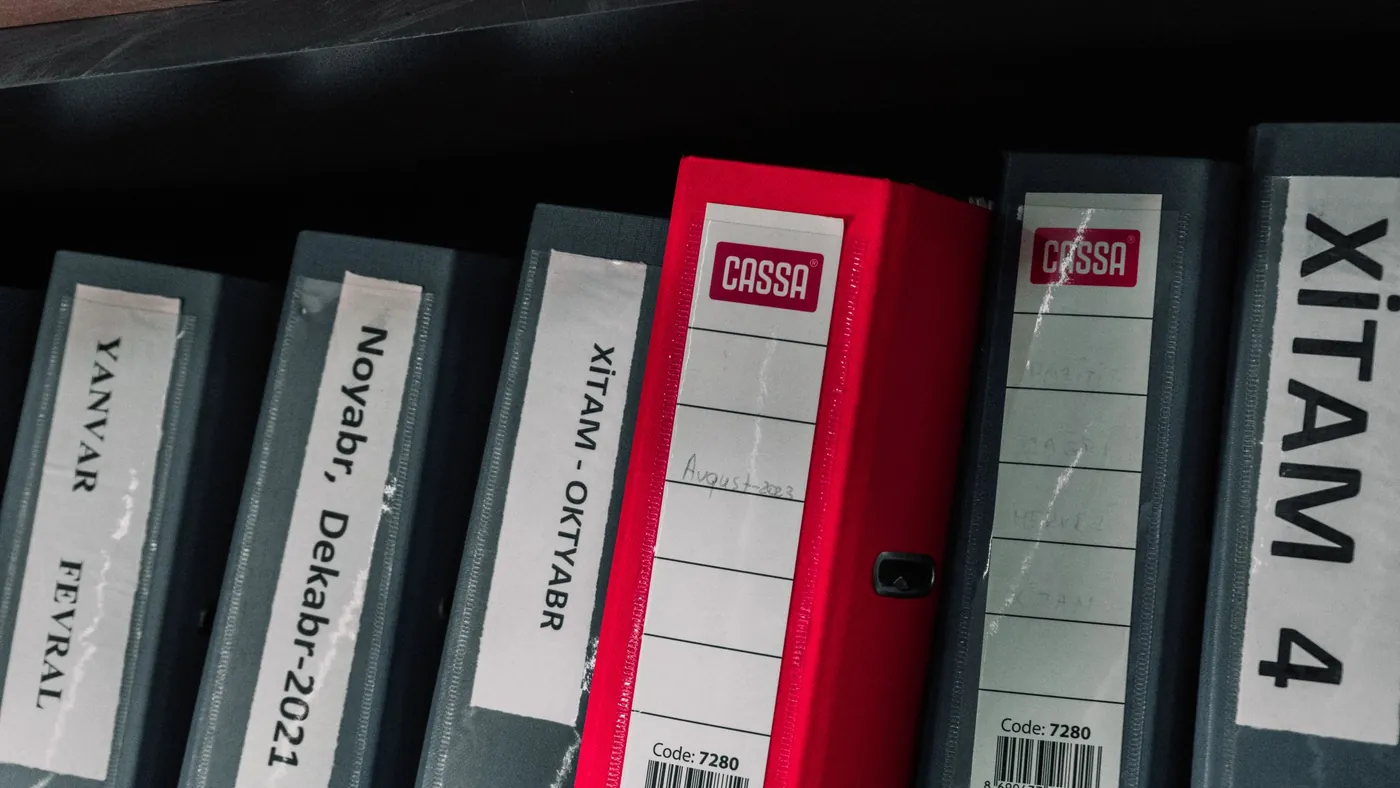 A row of labeled office binders on a shelf with one red binder standing out among grey ones